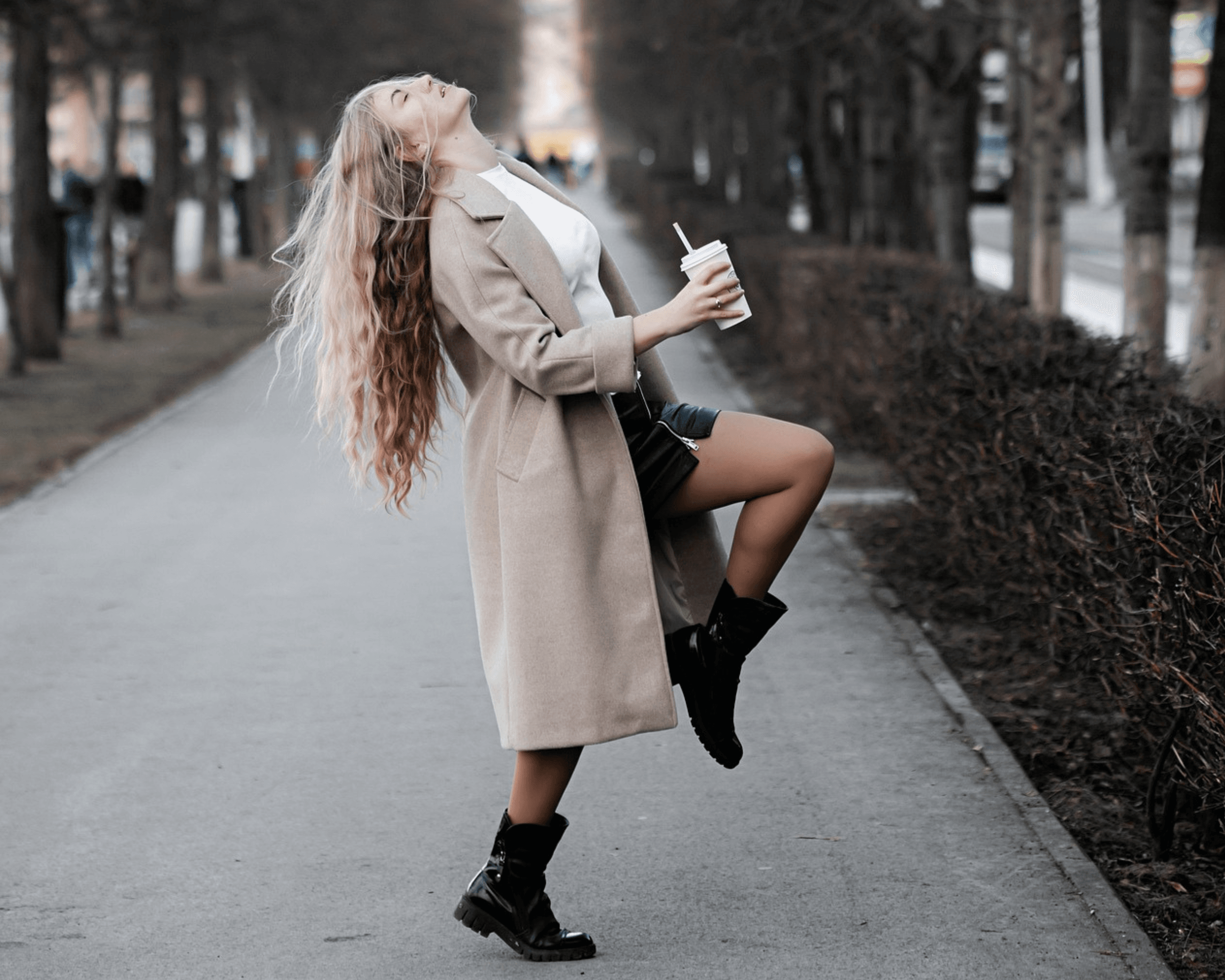 Woman wearing a beige wool coat, black boots, and leather shorts, tossing her hair back while holding a coffee – confident winter street style.