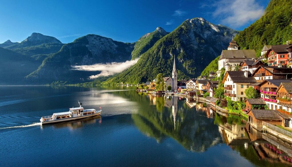 Scenic view of Hallstatt, Austria with colorful lakeside houses, a church tower, and a boat on the calm lake surrounded by the Alps