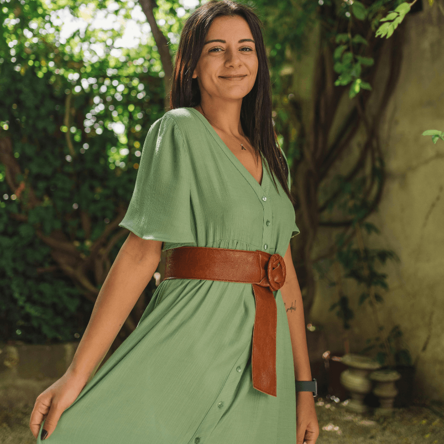 Woman wearing a green button-down dress with a wide brown leather belt, standing outdoors in sunlight.