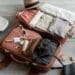 Flat lay of travel clothes, suitcase, and accessories on beige background – minimalist style