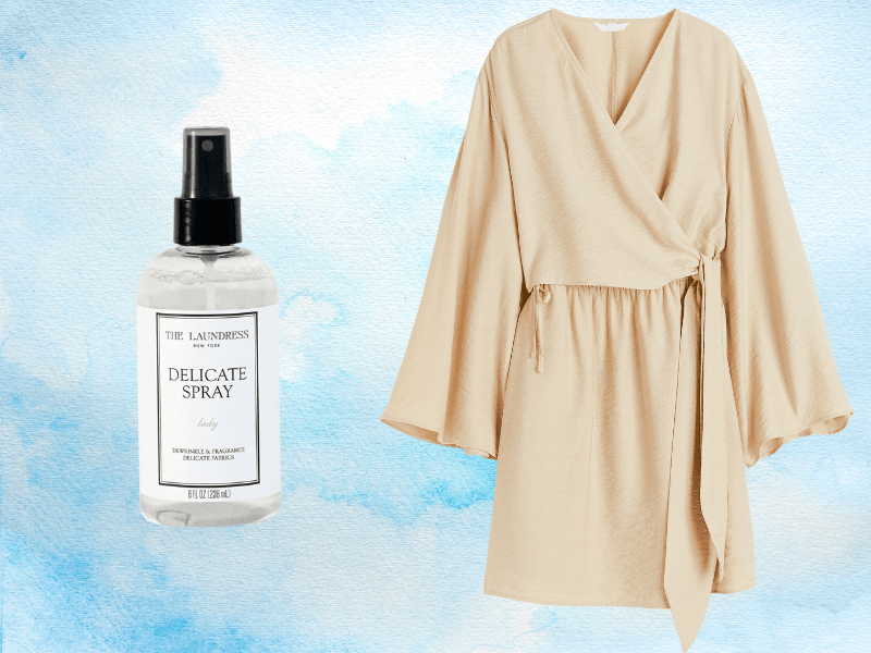 The Laundress Delicate Spray bottle next to a beige wrap dress on light blue watercolor background