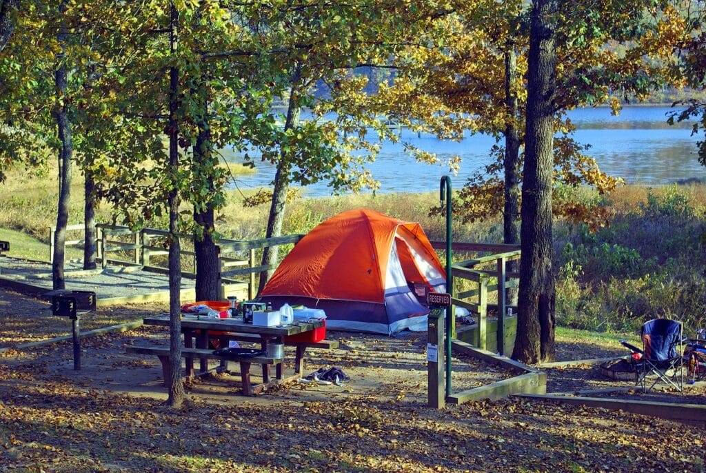 Scenic autumn campsite with an orange tent, picnic table full of gear, folding chairs, and lake view through trees.