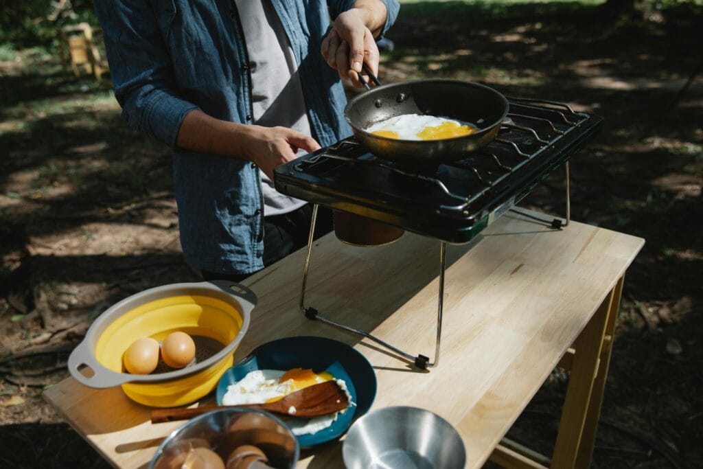 Person cooking eggs on a portable camping stove, with bowls and ingredients on a wooden table outdoors