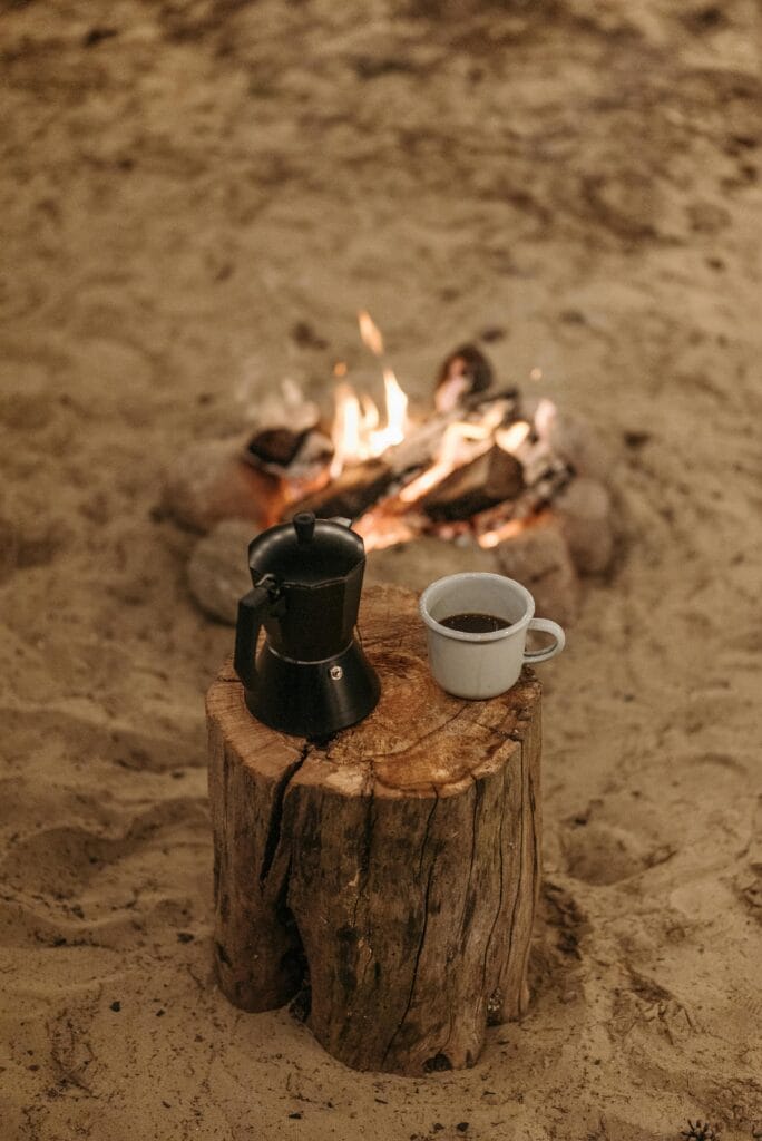 Black camping coffee pot and white enamel mug on a tree stump near a glowing campfire, creating a cozy outdoor morning scene