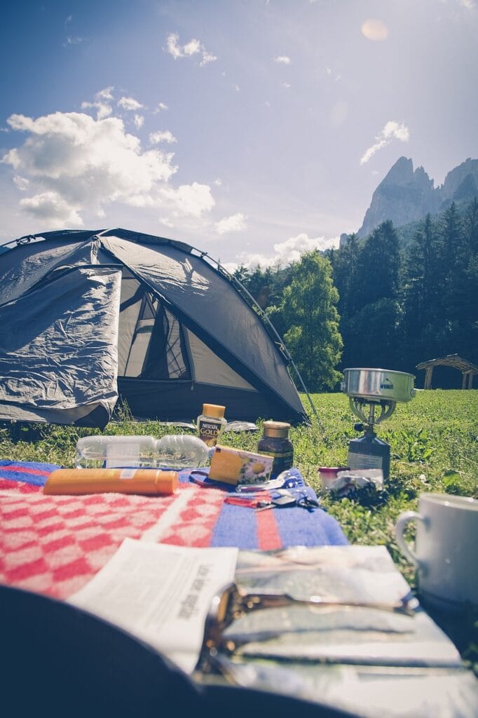 Morning camping setup with a tent, gas burner, coffee, sunscreen, and picnic items laid out on a colorful blanket in a mountain setting.