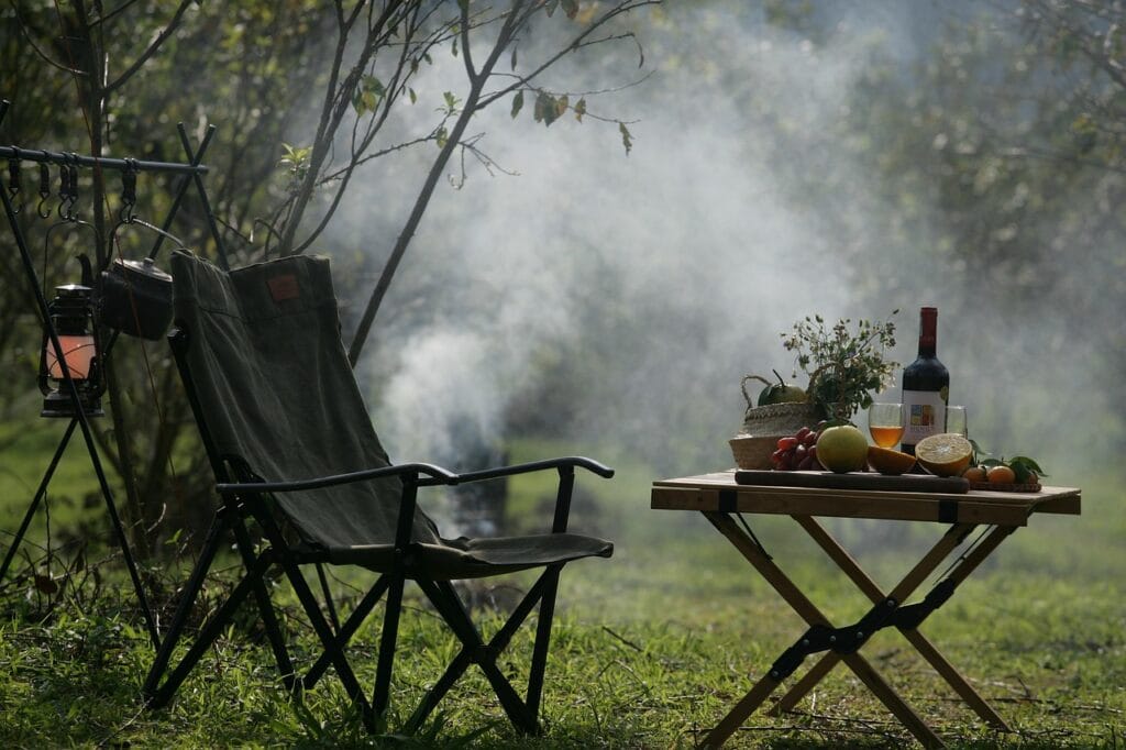 Foldable camping chair and wooden table with fruit, wine, and greenery, surrounded by soft smoke and trees — a peaceful and cozy outdoor setup.