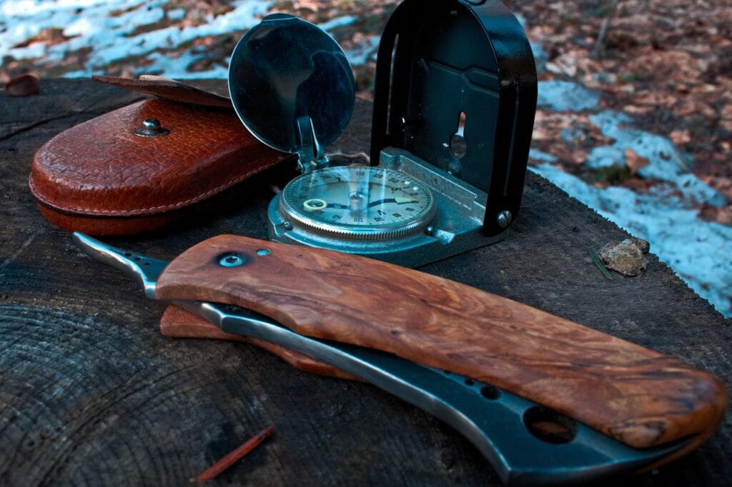 Close-up of a wooden-handled folding knife and an open metal compass placed on a tree stump in a forest setting.