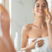 Woman smiling in front of mirror while cleansing her face with a sponge – fresh and radiant skincare morning routine