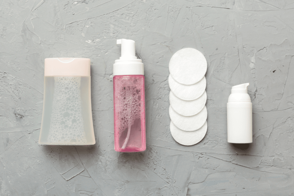 Flat lay of facial cleansing products – foaming cleanser, toner, cotton pads, and cream pump on gray background