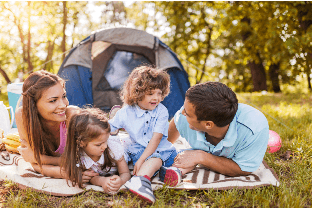 Family camping with young children outdoors, lying on a blanket in front of a tent and smiling together