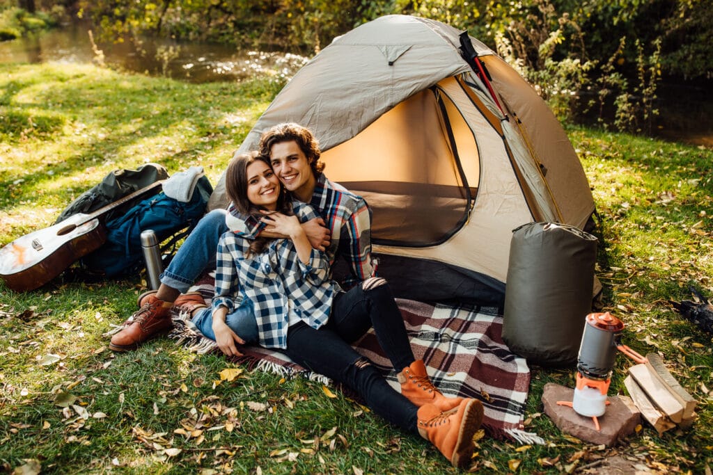 Smiling couple sitting on a blanket outside their tent, surrounded by camping gear, a guitar, and warm sunlight.