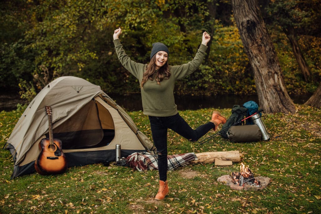Joyful young woman standing next to a tent and campfire in the forest, smiling and stretching with excitement.
