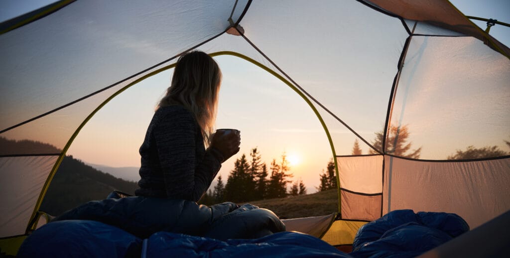 Woman sitting in a tent, drinking from a mug, watching the sunrise over forested mountains.