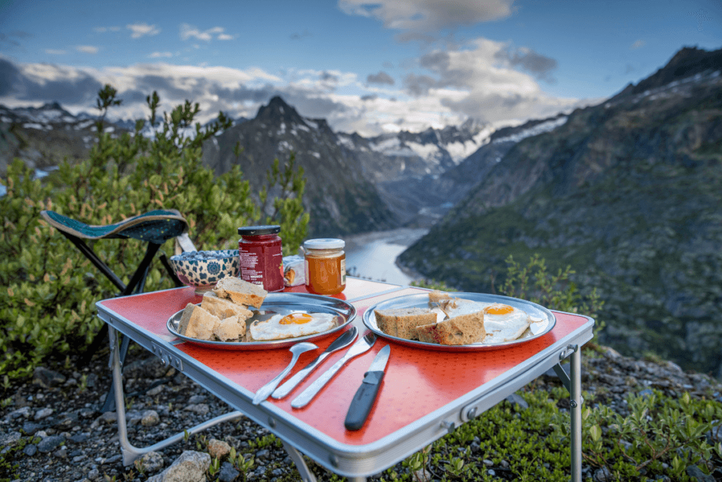 Outdoor camping breakfast setup with eggs, bread and jam on a table overlooking a mountain valley and river