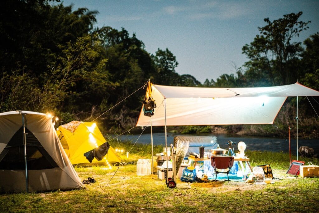 Nighttime camping setup with multiple tents, lights, tarp shelter, and full cooking and living gear beside a river.