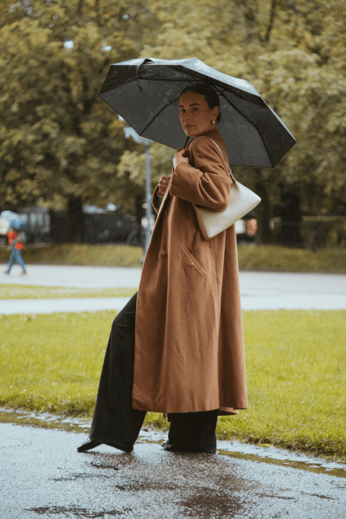 Woman wearing a long brown trench coat, black trousers, and holding a black umbrella on a rainy day in a park setting, carrying a white shoulder bag.