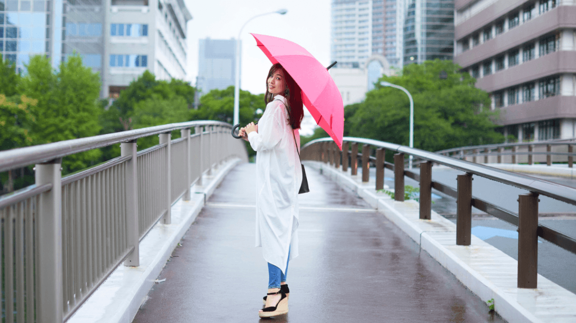 Woman walking on a city bridge wearing a long white raincoat, skinny jeans, wedge sandals, and holding a pink umbrella on a rainy summer day.