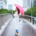 Woman walking on a city bridge wearing a long white raincoat, skinny jeans, wedge sandals, and holding a pink umbrella on a rainy summer day.
