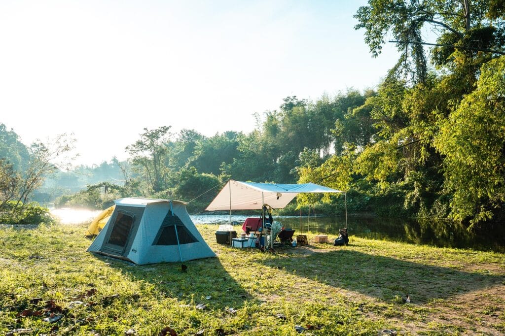 Fully set up camping spot by a river, with a large tent, shaded tarp area, and supplies — surrounded by greenery and morning sunlight.