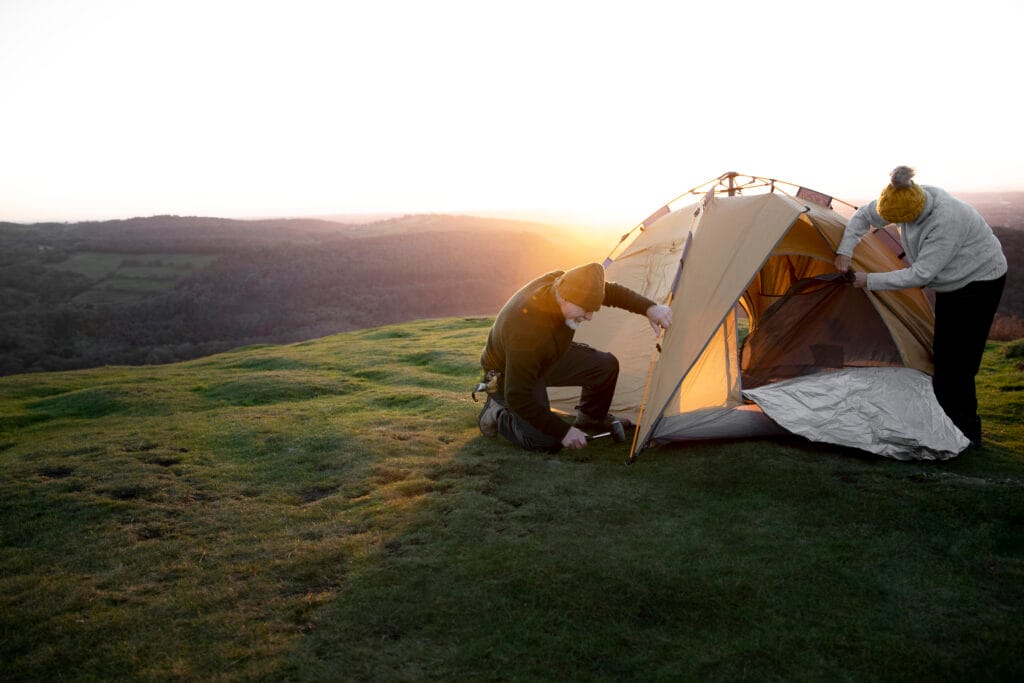 Two people setting up a tent on a grassy hill at sunset, with scenic forested hills in the background.