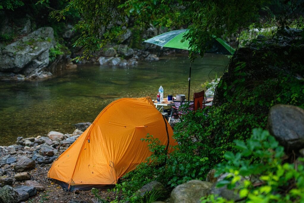 A bright orange tent set up beside a quiet river, with a camp table and umbrella under light rain, surrounded by rocks and forest greenery.