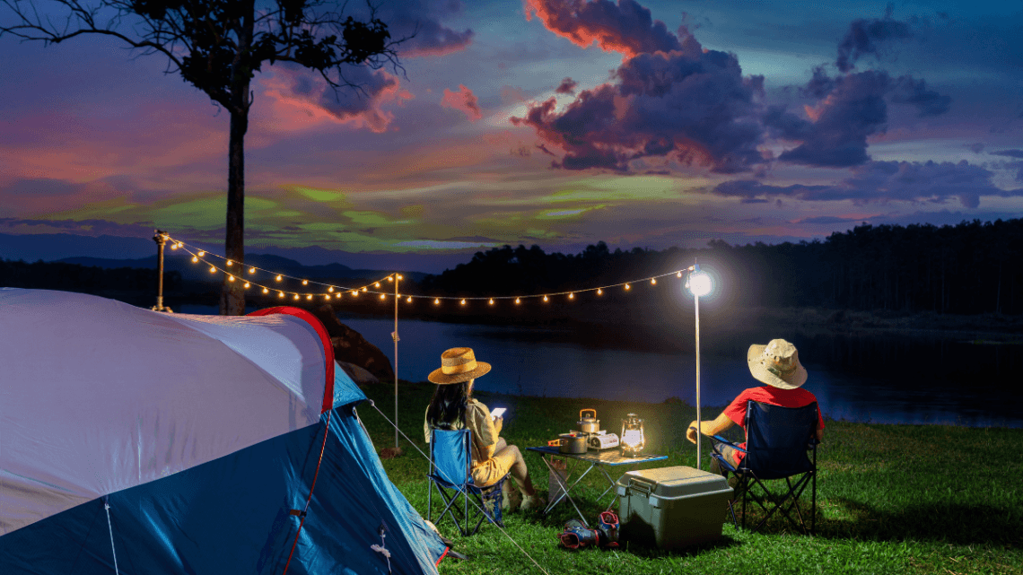 Two people sitting by a tent at sunset, camping by a lake with string lights and cooking gear.