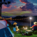 Two people sitting by a tent at sunset, camping by a lake with string lights and cooking gear.