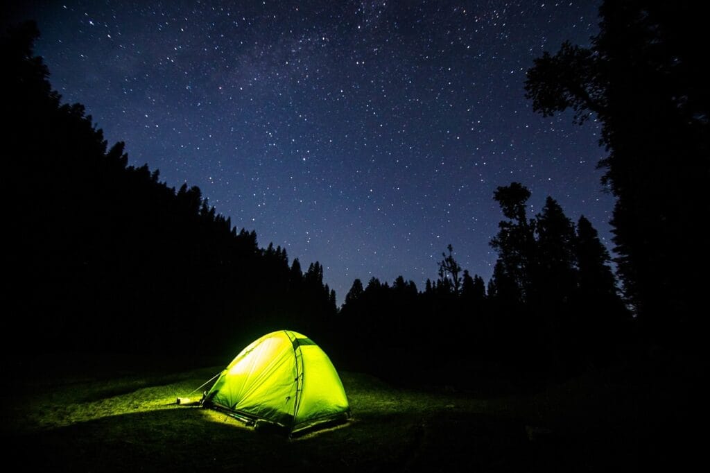 Glowing tent at night under a starry sky surrounded by forest silhouettes in a peaceful camping scene
