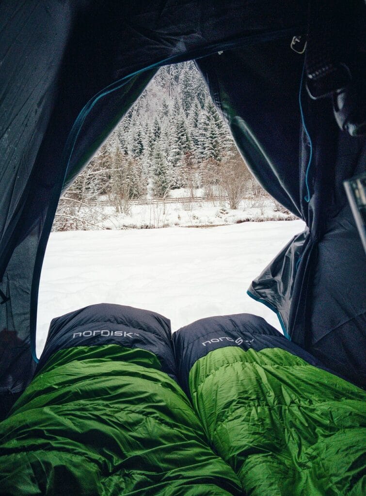 Two winter sleeping bags inside a tent, looking out at a snow-covered forest landscape.