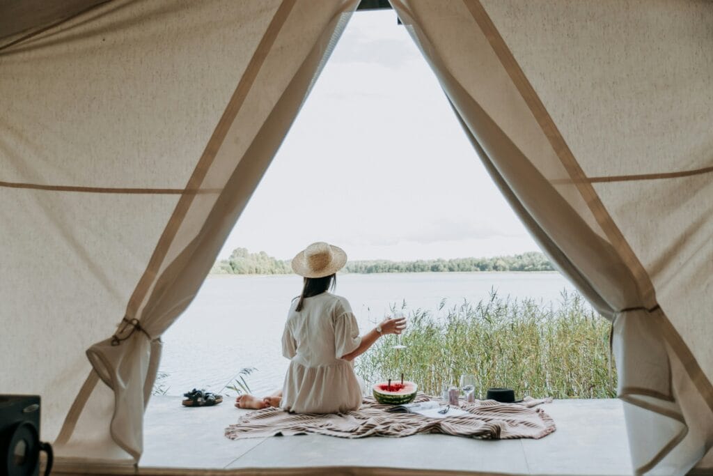 Woman sitting inside a tent, looking at a lake, enjoying a quiet morning with a glass of water and fruit