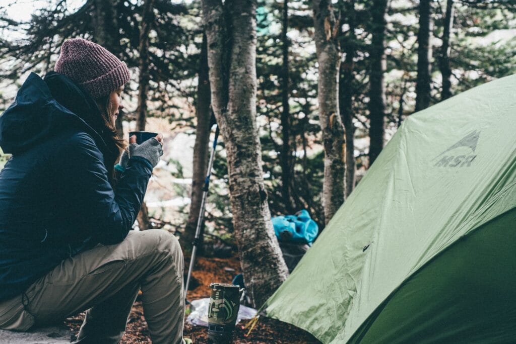 Woman dressed warmly, sitting by a tent in the forest and drinking coffee in the morning cold air