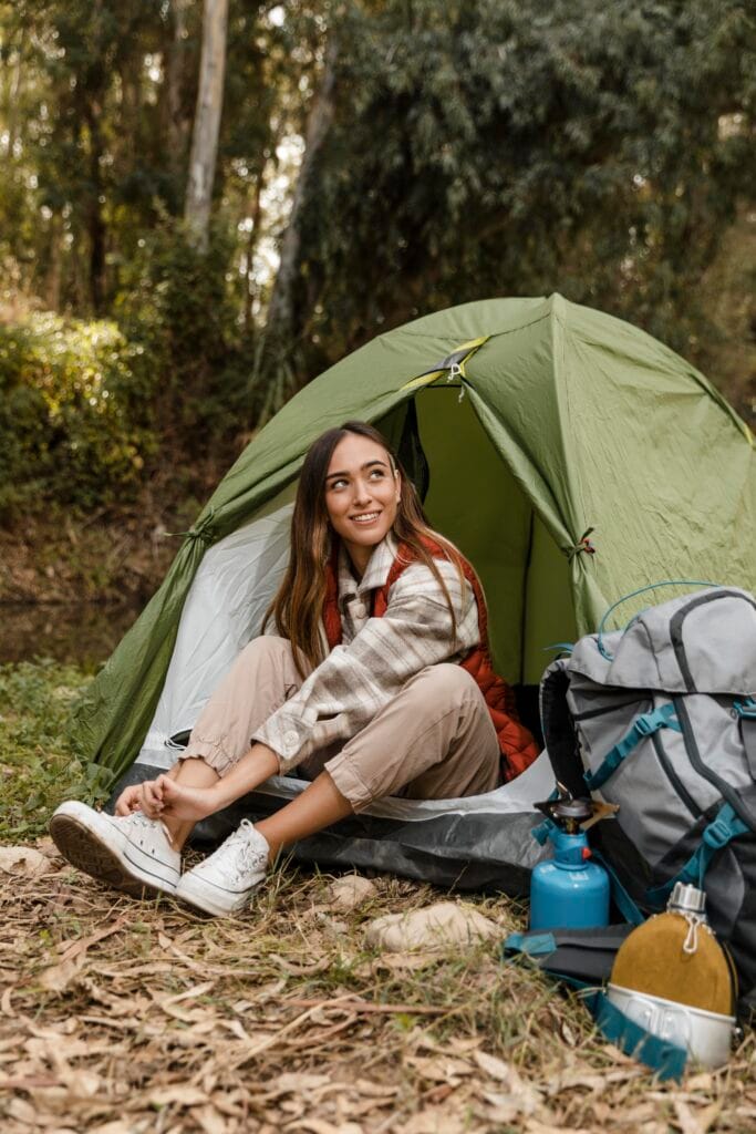 Young woman sitting at the entrance of a tent in the forest, smiling while putting on her shoes, with camping gear beside her