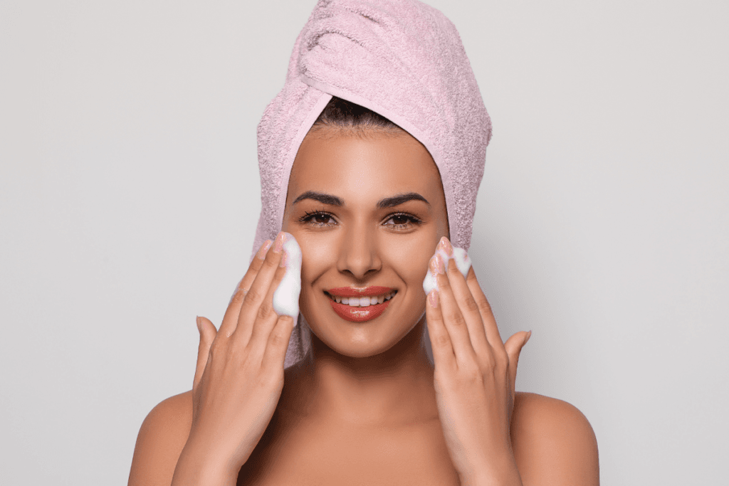 Smiling woman with a towel on her head applying foam cleanser on her face – joyful skincare moment