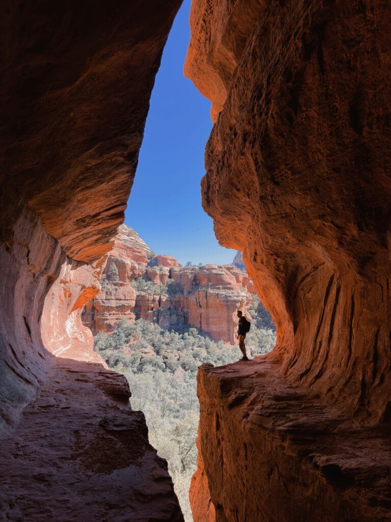Hiker standing inside Subway Cave on Boynton Canyon Trail in Sedona, Arizona, with red rock cliffs and blue sky in the background