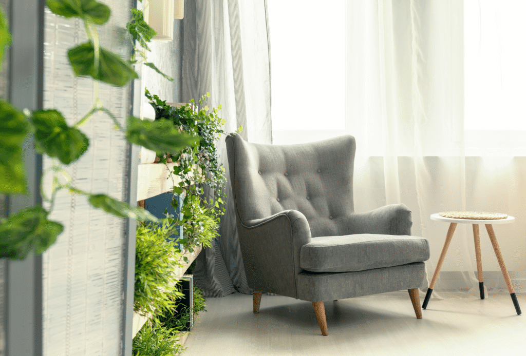 Cozy reading nook with a gray tufted armchair, small wooden side table, and vibrant indoor plants near a sunlit window with sheer curtainsCozy reading nook with a gray tufted armchair, small wooden side table, and vibrant indoor plants near a sunlit window with sheer curtains